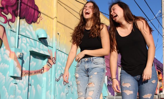 Dua wanita memakai tanktop warna hitam dan celana jeans biru sedang tertawa